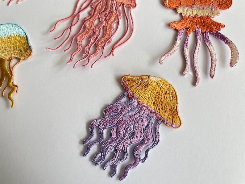 May include: Embroidered jellyfish patches in shades of yellow, orange, pink, and purple. The patches feature detailed stitching, with flowing tentacles and a rounded bell shape. These patches are ideal for adding a unique touch to clothing or accessories.