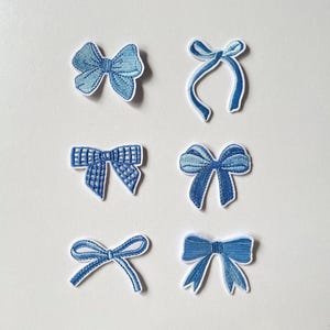 May include: Six embroidered patches in various shades of blue, each shaped like a decorative bow. The designs include a solid textured bow, a thin ribbon-style bow, a checkered pattern bow, a classic tied bow, a slender looped bow, and a wide bow.