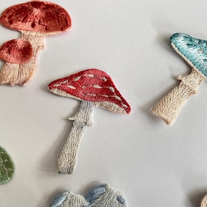 May include: Embroidered mushroom patches in various colors, including red, pink, and blue. The patches feature detailed stitching and are arranged on a white background. These patches are ideal for adding a whimsical touch to clothing or accessories.