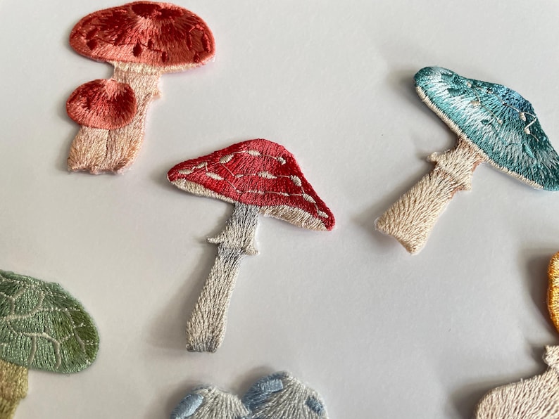 May include: Embroidered mushroom patches in various colors, including red, pink, and blue. The patches feature detailed stitching and are arranged on a white background. These patches are ideal for adding a whimsical touch to clothing or accessories.