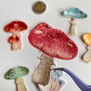 May include: Embroidered mushroom patches in various colors, including red, green, and blue. The patches have detailed stitching and are in different sizes. A pair of tweezers holds one patch, and a coin is also visible.