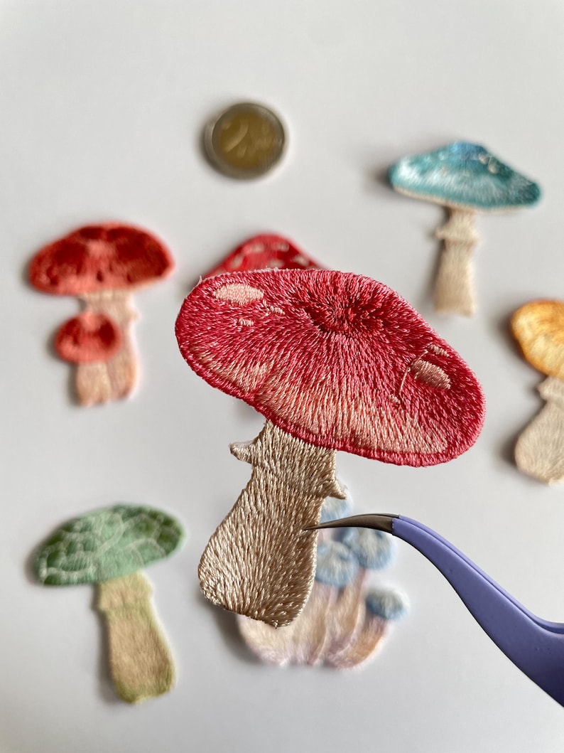 May include: Embroidered mushroom patches in various colors, including red, green, and blue. The patches have detailed stitching and are in different sizes. A pair of tweezers holds one patch, and a coin is also visible.
