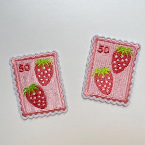 May include: Two embroidered patches with a strawberry design. Each patch has a pink background with a red strawberry and green leaves. The number "50" is printed at the top. The patches have a scalloped edge.