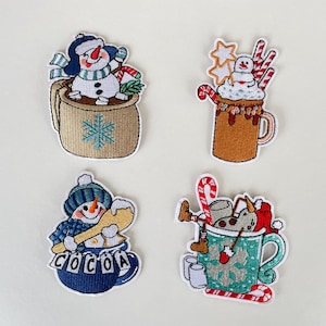 Op de afbeelding: Vier geborduurde patches met sneeuwmannen en mokken. Een sneeuwman zit in een beige mok met een sneeuwvlok, een andere in een bruine mok met slagroom en zuurstokken, een derde met het woord "COCOA" op een blauwe mok en de laatste in een turquoise mok.