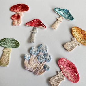 May include: Embroidered mushroom patches in various colors, including red, green, blue, and orange. The patches feature detailed stitching and are arranged on a white surface. These patches are ideal for adding a whimsical touch to clothing or accessories.