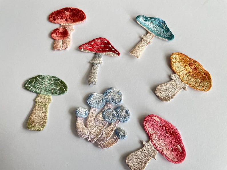 May include: Embroidered mushroom patches in various colors, including red, green, blue, and orange. The patches feature detailed stitching and are arranged on a white surface. These patches are ideal for adding a whimsical touch to clothing or accessories.