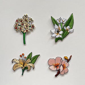 Pode incluir: Quatro patches florais bordados. Um patch apresenta um grupo de pequenas flores creme com detalhes vermelhos e um caule verde. Outro mostra flores brancas com folhas verdes. Um terceiro patch retrata um lírio amarelo. O último mostra flores cor-de-rosa.