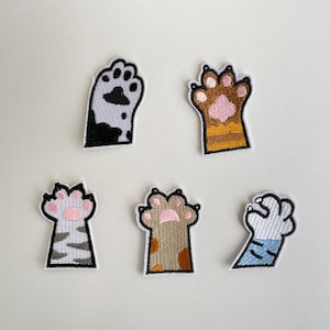 Può includere: Cinque toppe ricamate a forma di zampa di gatto in vari colori. Le toppe presentano disegni neri e bianchi, marroni e gialli, grigi e rosa, marroni e arancioni e blu e bianchi. Ogni toppa ha un bordo nero.