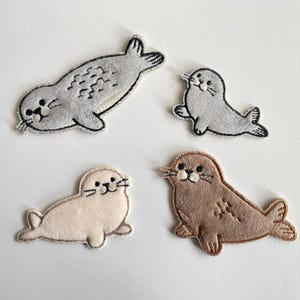 Op de afbeelding: Vier geborduurde zeehondenpatches in verschillende kleuren: grijs, lichtgrijs, beige en bruin. Elke patch heeft een lachend zeehondenontwerp met zwarte ogen en snorharen. De zeehonden staan in verschillende poses.