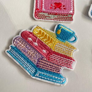 May include: Embroidered patches depicting a stack of colourful books and a blue teacup. The books are in shades of blue, pink, yellow, and purple, with white stitching. The teacup has yellow liquid. The patches have a white border.