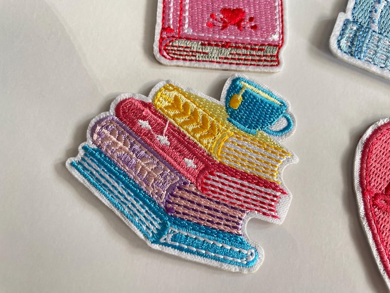 May include: Embroidered patches depicting a stack of colourful books and a blue teacup. The books are in shades of blue, pink, yellow, and purple, with white stitching. The teacup has yellow liquid. The patches have a white border.