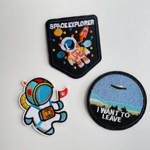 May include: Three embroidered space-themed patches. One is a black shield shape with an astronaut and the words "SPACE EXPLORER." Another is a round patch with a UFO and the words "I WANT TO LEAVE." The third patch shows an astronaut holding a flag.