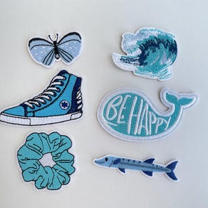 May include: A collection of embroidered patches in shades of blue. Designs include a butterfly, a high-top sneaker, a wave, a whale with "BE HAPPY" text, a scrunchie, and a fish. Each patch has a white border.