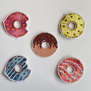 May include: Five embroidered donut patches in various colors. One pink donut has a bite taken out of it. Other donuts are brown with chocolate frosting, blue with white stripes, yellow with white frosting, and pink with sprinkles.
