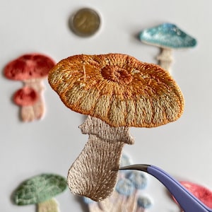 May include: Embroidered mushroom patches in various colors, including orange, red, green, and blue. The patches have detailed stitching and are arranged on a white surface. A pair of tweezers is holding one of the patches.