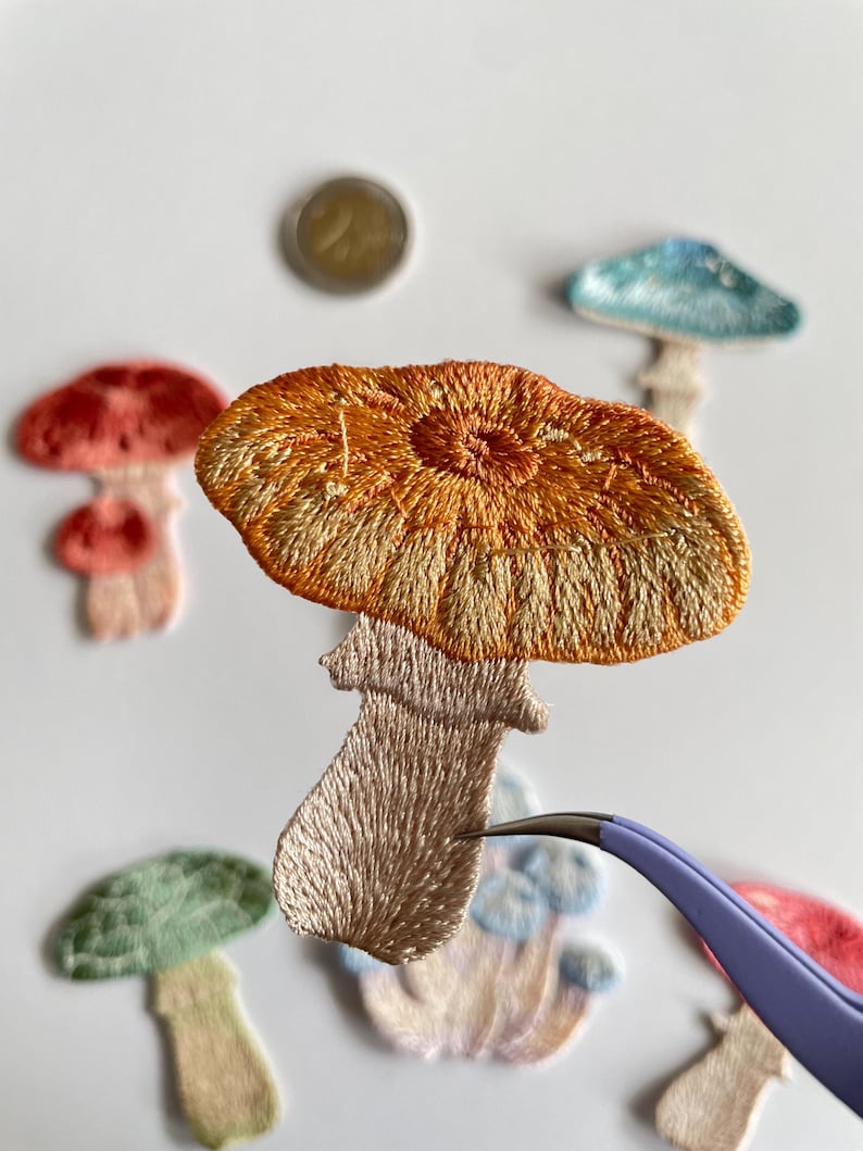 May include: Embroidered mushroom patches in various colors, including orange, red, green, and blue. The patches have detailed stitching and are arranged on a white surface. A pair of tweezers is holding one of the patches.
