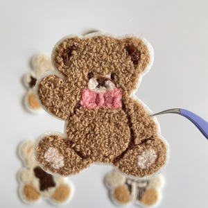 Pode incluir: Um patch de urso de pel&uacute;cia marrom com uma gravata borboleta rosa e acabamento branco. O urso tem uma apar&ecirc;ncia texturizada e difusa. Patches de urso adicionais s&atilde;o vis&iacute;veis no fundo.