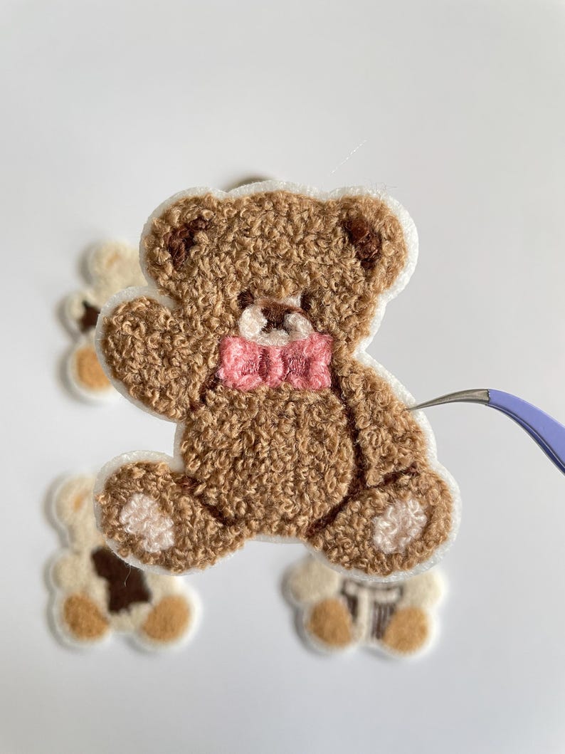 Pode incluir: Um patch de urso de pel&uacute;cia marrom com uma gravata borboleta rosa e acabamento branco. O urso tem uma apar&ecirc;ncia texturizada e difusa. Patches de urso adicionais s&atilde;o vis&iacute;veis no fundo.