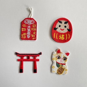 Könnte beinhalten: Vier bestickte Aufnäher mit japanischen Motiven: ein rotes, rechteckiges Omamori-Amulett mit weißem Text, eine runde rote Daruma-Puppe mit schwarzen und gelben Details, ein rot-schwarzes Torii-Tor und eine weiße Winkekatze mit Goldakzenten.