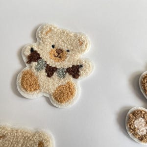 Pode incluir: Close-up de v&aacute;rios patches de ursinhos de pel&uacute;cia. Os patches s&atilde;o feitos de um material macio e texturizado nas cores creme, castanho e bege. Um urso tem uma pequena guirlanda em volta do pesco&ccedil;o. Estes patches s&atilde;o ideais para adicionar um toque fofo a roupas ou acess&oacute;rios.