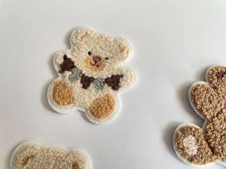 Pode incluir: Close-up de v&aacute;rios patches de ursinhos de pel&uacute;cia. Os patches s&atilde;o feitos de um material macio e texturizado nas cores creme, castanho e bege. Um urso tem uma pequena guirlanda em volta do pesco&ccedil;o. Estes patches s&atilde;o ideais para adicionar um toque fofo a roupas ou acess&oacute;rios.