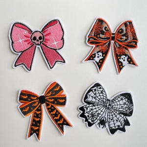 Könnte beinhalten: Vier gestickte Halloween-Themen-Schleifen-Patches. Zu den Designs gehören eine rosa Schleife mit einem Totenkopf, eine orangefarbene Schleife mit Geistern, eine orange-schwarze Schleife mit Fledermäusen und eine schwarz-weiße Schleife mit Spinnennetz und Spinne.