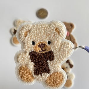 Pode incluir: Close-up de um patch de urso de pel&uacute;cia bege com uma gravata borboleta e detalhes marrons. O urso tem olhos pretos e &eacute; cercado por uma borda branca. Outros patches semelhantes s&atilde;o ligeiramente vis&iacute;veis no fundo.