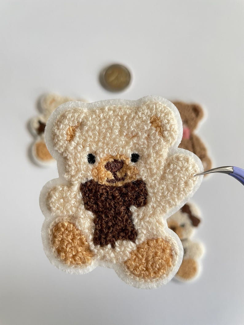 Pode incluir: Close-up de um patch de urso de pel&uacute;cia bege com uma gravata borboleta e detalhes marrons. O urso tem olhos pretos e &eacute; cercado por uma borda branca. Outros patches semelhantes s&atilde;o ligeiramente vis&iacute;veis no fundo.