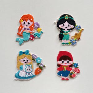 May include: Four embroidered patches featuring cartoon characters: a mermaid with orange hair and a teal tail, a princess in green, Alice in a blue dress, and Little Red Riding Hood. Each patch has a unique floral or thematic accent.
