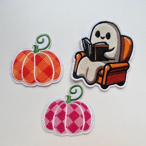 May include: Three embroidered patches: two pumpkins and a ghost reading a book. The pumpkins are orange and pink with a plaid pattern. The ghost is white, sitting in an orange armchair, reading a black book.
