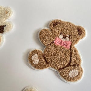 Pode incluir: Close-up de v&aacute;rios patches de ursinhos de pel&uacute;cia. Os patches s&atilde;o castanhos e creme com uma apar&ecirc;ncia texturizada e difusa. Um urso tem uma gravata borboleta rosa. Os patches est&atilde;o em um fundo branco.