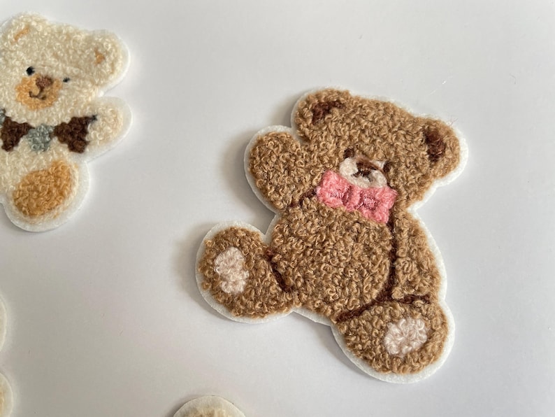 Pode incluir: Close-up de v&aacute;rios patches de ursinhos de pel&uacute;cia. Os patches s&atilde;o castanhos e creme com uma apar&ecirc;ncia texturizada e difusa. Um urso tem uma gravata borboleta rosa. Os patches est&atilde;o em um fundo branco.