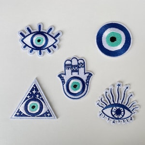 Könnte beinhalten: Eine Sammlung von fünf gestickten Aufnähern mit dem Symbol des bösen Blicks. Die Aufnäher haben verschiedene Formen, darunter ein Auge, ein Kreis, ein Dreieck, eine Hand und ein Auge mit Strahlen. Die Aufnäher sind hauptsächlich blau, weiß und türkis.