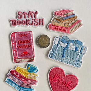 May include: Embroidered patches with a book theme. The patches feature stacks of books in different colours, a heart with the word "Book", and the phrase "Stay Bookish". A coin is also present.