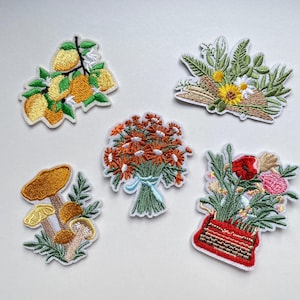 Könnte beinhalten: Gestickte Aufnäher mit floralen und botanischen Motiven. Enthält einen Zitronenzweig, einen Strauß orangefarbener Blumen, eine Pilzgruppe, ein Blumenarrangement und einen Strauß mit einer Sonnenblume. Jeder Aufnäher hat einen weißen Rand.