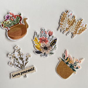 Puede incluir: Parches bordados con diseños florales. Incluye una tetera con flores, un ramo, espigas de trigo, un arreglo floral con el texto "Love yourself" y una cesta de flores. Los parches tienen bordes blancos.