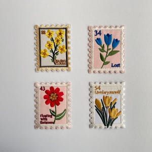 Puede incluir: Cuatro parches bordados rectangulares con forma de sello postal. Arriba a la izquierda, flores amarillas con el texto "CHASING CLOUDS". Arriba a la derecha, tulipanes azules y la palabra "Lost". Abajo a la izquierda, una flor roja y "Floating with the breeze". Abajo a la derecha, azafranes amarillos y "Live for yourself".