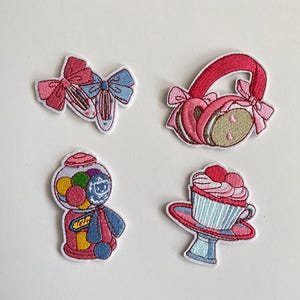 May include: Embroidered patches featuring a ballet shoe with pink and blue bows, pink earmuffs with bows, a gumball machine, and a cupcake with pink frosting and cherries. These patches are in shades of pink, blue, and white.