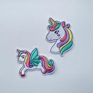 Puede incluir: Dos parches bordados de unicornios. Uno representa una cabeza de unicornio con un cuerno dorado, melena rosa y detalles morados y amarillos. El otro muestra un unicornio con alas, un cuerno dorado y una melena y cola arcoíris.