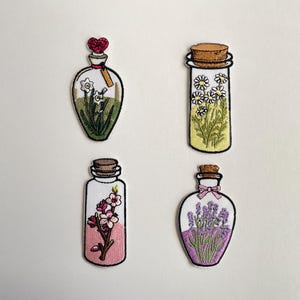Könnte beinhalten: Vier bestickte Aufnäher in Form von Glasflaschen. Einer enthält weiße Blumen mit einem roten Herzverschluss, einer weiße Gänseblümchen mit Korken, einer rosa Blüten in einer klaren Flüssigkeit und einer violetten Lavendel mit einer rosa Schleife.