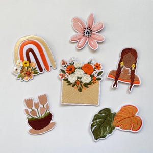 Op de afbeelding: Een verzameling van zes geborduurde patches met een regenboog met bloemen, een roze bloem, een boeket oranje en witte bloemen in een envelop, een persoon met gevlochten haar, een potplant met tulpen en twee grote tropische bladeren.