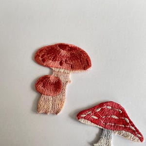 May include: Embroidered mushroom patches in shades of red, pink, and beige. The patches feature detailed stitching, with one mushroom having a textured cap and another with white spots. These patches are ideal for adding a whimsical touch to clothing or accessories.