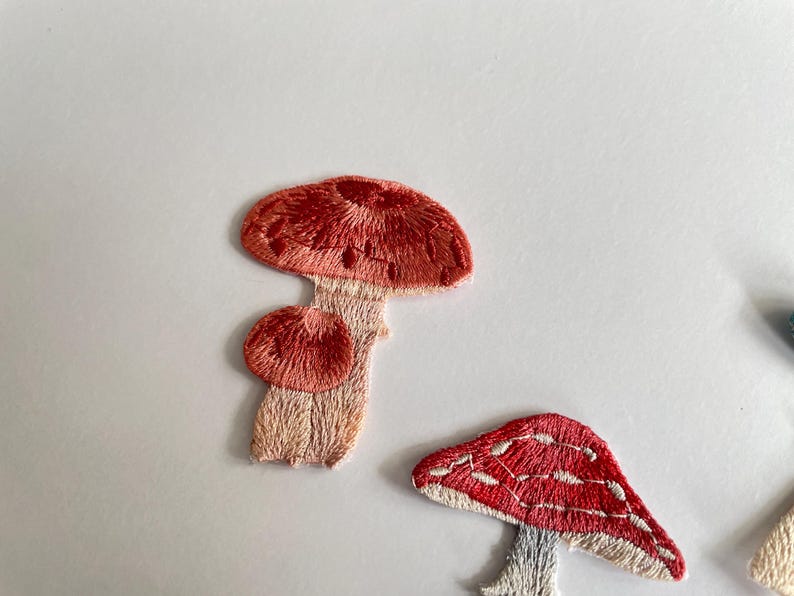 May include: Embroidered mushroom patches in shades of red, pink, and beige. The patches feature detailed stitching, with one mushroom having a textured cap and another with white spots. These patches are ideal for adding a whimsical touch to clothing or accessories.