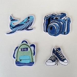Könnte beinhalten: Vier gestickte Aufnäher: ein blauer Wal, eine blaue Kamera, ein grüner Rucksack mit dem Text "BYE" und ein Paar blaue Turnschuhe. Jeder Aufnäher hat einen weißen Rand auf weißem Hintergrund.