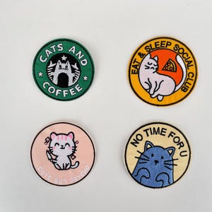 Könnte beinhalten: Vier runde, gestickte Aufnäher mit Katzenmotiven. Ein grüner Aufnäher trägt die Aufschrift "Cats and Coffee". Ein anderer zeigt eine Katze mit Pizza und dem Text "Eat & Sleep Social Club". Der dritte Aufnäher trägt die Aufschrift "Cute But Psycho". Der letzte Aufnäher trägt die Aufschrift "No Time For U."