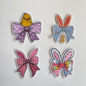 Könnte beinhalten: Vier gestickte Aufnäher mit Ostermotiven. Einer zeigt ein gelbes Küken über einer lila Schleife mit Punkten. Andere zeigen Hasenohren mit Schleifen in Rosa, Blau und gemusterten Designs. Ideal, um Kleidung oder Accessoires eine festliche Note zu verleihen.