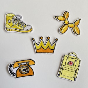 Könnte beinhalten: Eine Sammlung gelber, gestickter Aufnäher mit Motiven wie einem High-Top-Sneaker mit Stern, einem Ballonhund, einer Krone, einem Wählscheibentelefon und einem Rucksack. Jeder Aufnäher hat einen weißen Rand.