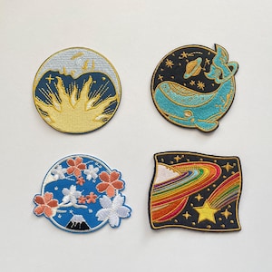 Könnte beinhalten: Vier gestickte Aufnäher mit himmlischen und Naturthemen. Designs umfassen eine Sonne, einen Wal im Weltraum, Kirschblüten mit einem Berg und einen Regenbogen mit einem Stern. Jeder Aufnäher hat eine andere Form und Farbgebung.