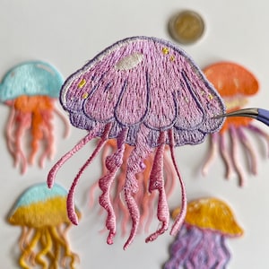 May include: Embroidered jellyfish patches in various colors, including purple, orange, yellow, and blue. The patches feature detailed stitching and flowing tentacles. A coin is visible in the background. These patches are suitable for clothing or craft projects.