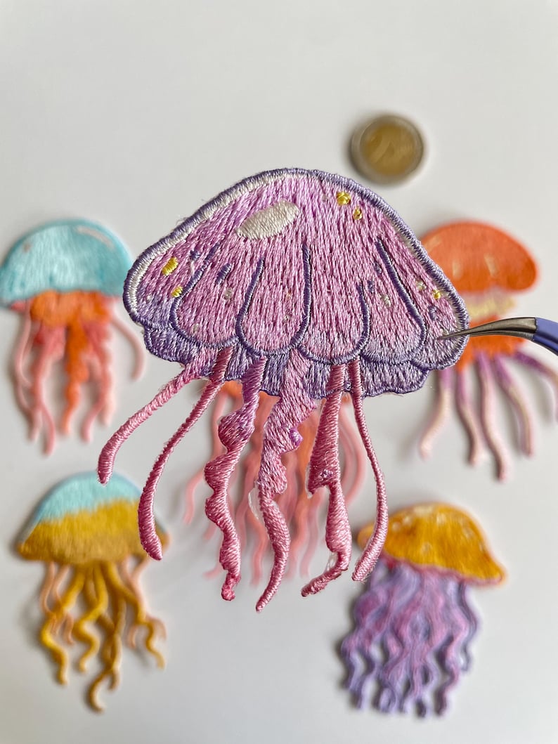 May include: Embroidered jellyfish patches in various colors, including purple, orange, yellow, and blue. The patches feature detailed stitching and flowing tentacles. A coin is visible in the background. These patches are suitable for clothing or craft projects.